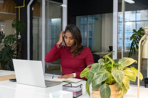 Indian woman working at modern office desk holding phone checking laptop and notebook