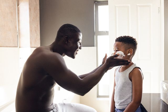 This image captures a joyful moment of a father and son bonding in the bathroom. The father is playfully applying shaving foam to his son's face, creating a fun and loving atmosphere. Ideal for use in articles or advertisements about parenting, family activities, morning routines, or personal hygiene products. It can also be used to illustrate the importance of quality family time and father-son relationships.