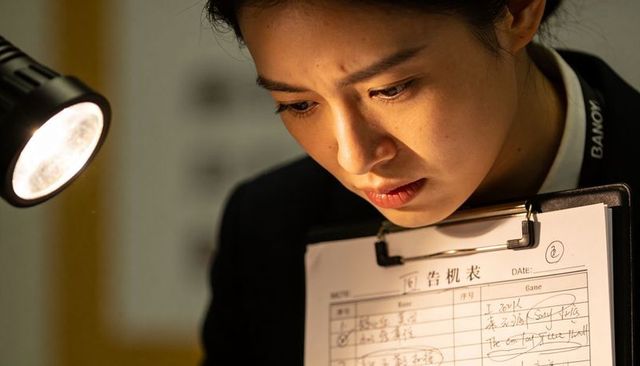 Asian inspector leaning over clipboard reading document under desk lamp spotlight