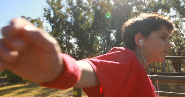 Boy running in park with focused expression, wearing red wristband and earphones. Sunlight filtering through trees in background. Great for concepts of active lifestyle, exercise, health, and determination. Suitable for fitness promotions, healthy lifestyle campaigns, and outdoor activity advertisements.