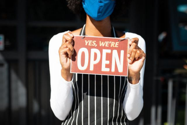 African american female barista wearing face mask and apron holding open sign at work. business and work during coronavirus covid 19 pandemic.