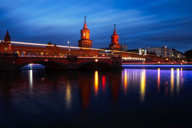 Illuminated bridge spanning serene river at night in Berlin. Twilight sky adds dramatic effect with the city lights reflecting in the water. Suitable for travel brochures, blog articles on urban destinations, or tourism-related promotional material.