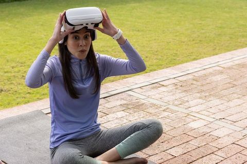Asian woman experimenting with vr headset while sitting cross-legged on outdoor yoga mat