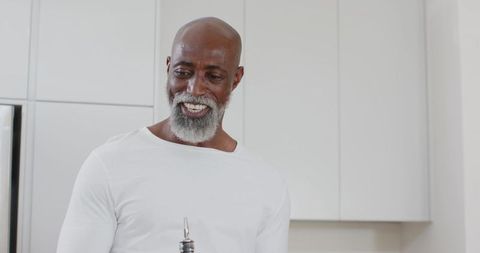 Mature african man smiling and holding oil bottle with pour spout in modern white kitchen