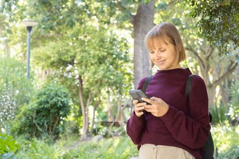 Young woman smiling while texting on smartphone in sunlit park wearing maroon sweater