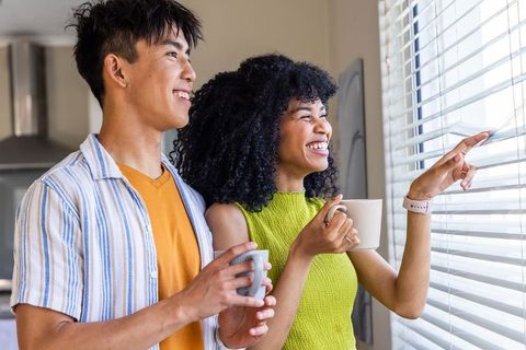 Multicultural couple enjoying morning coffee by window blinds, smiling and pointing