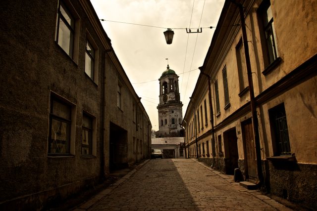 This image shows a vintage cobblestone street lined with old buildings and culminates with a historical church tower in the background. The scene, bathed in muted and warm tones, suggests an atmosphere of nostalgia and architectural heritage. Ideal for use in travel brochures, historical websites, cultural documentaries, or any content that seeks to evoke a sense of old-world charm and historical depth.