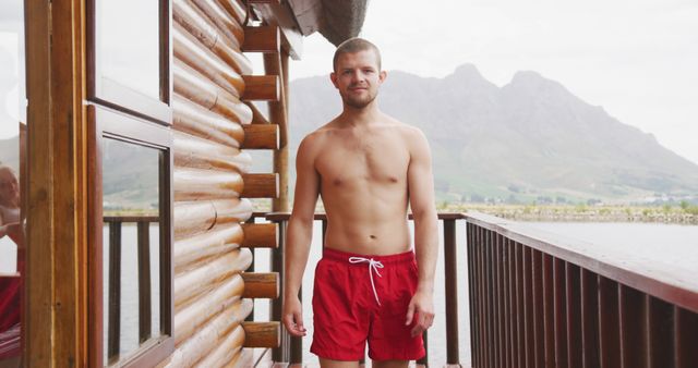 Young man standing on the balcony of a rustic wooden cabin overlooking a serene lake and distant mountains, popular for promoting vacation rentals, lifestyle blogs, outdoor getaways, and summer travel destinations.