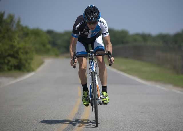 Cyclist in aerodynamic position riding a bicycle on an open rural road. Ideal for use in publications related to competitive cycling, outdoor sports, fitness, endurance training, athletic equipment, and healthy lifestyle.