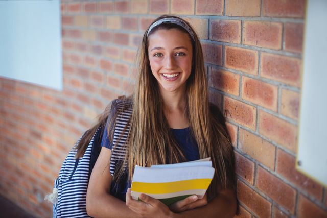Happy schoolgirl holding books and standing near brick wall in school
