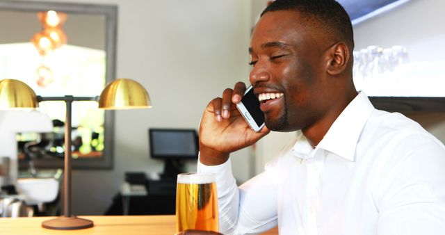 Image of a smiling businessman enjoying a pint of beer at a bar while talking on his phone. This can be used to illustrate concepts of work-life balance, relaxation, socializing, communication, and modern lifestyle. Ideal for use in articles or advertisements related to business, leisure, and social activities.