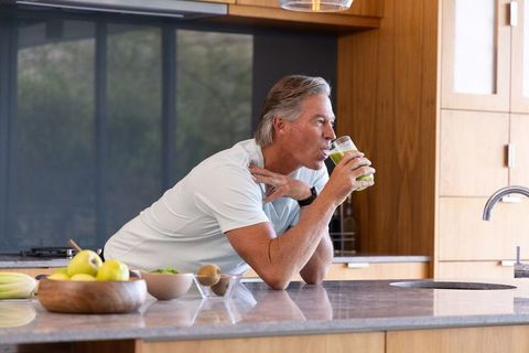 Mature man drinking green smoothie in modern kitchen leaning on marble island