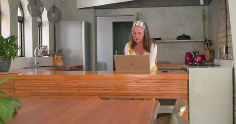 Mature woman in yellow apron working on laptop at kitchen island in sunlit home office