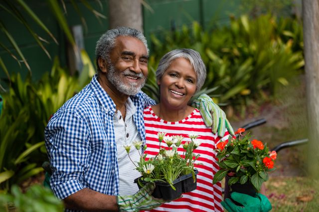 Senior couple standing together in their backyard, both smiling and holding plants. The lush greenery and vibrant flowers around them create a serene and joyful atmosphere. Suitable for promoting healthy lifestyles, outdoor activities, and family bonding. Ideal for use in retirement community advertisements, gardening promotions, and wellness blogs.