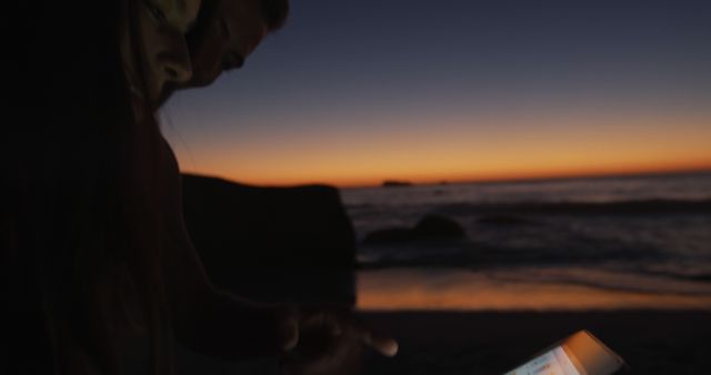 Couple engrossed with smartphone on a beach as sun sets, producing serene evening scene. Great for concepts of romance, technology, social media interaction, and tranquil coastal moments.