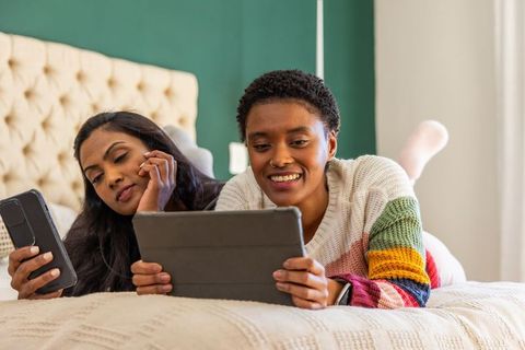 Black woman and south asian woman lying on bed, enjoying tablet together, cozy lifestyle