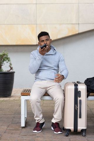 African american traveler sitting on bench talking on smartphone with beige suitcase waiting