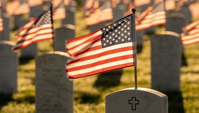 American flag fluttering over veteran grave with cross in sunlit cemetery at sunrise