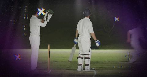 Night cricket batsman standing near stumps under floodlights holding bat with pads and helmet