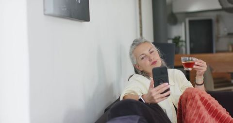 Relaxed senior woman reclining on sofa, holding smartphone and tea, cozy home moment