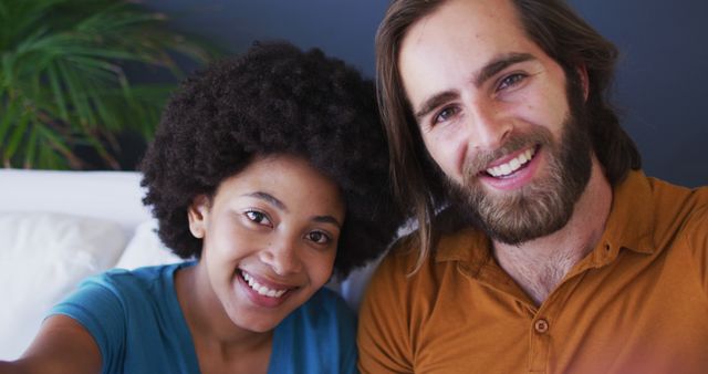 Happy interracial couple taking selfie indoors, showing joy and togetherness. Great for use in relationship and family themes, advertisements for lifestyle or home decor products, and articles promoting diversity and social integration.