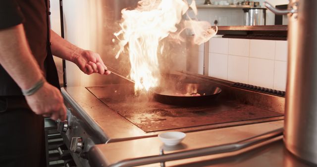 Captures professional chef hand holding pan, flambéing on restaurant stove. Ideal for articles about culinary arts, cooking techniques, restaurant industry, chef training programs, gourmet cuisine advertisement.