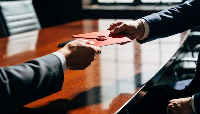 Executives exchanging wax-sealed red envelope over polished mahogany conference table, boardroom
