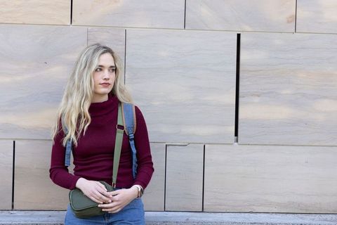 Young woman leaning against sandstone wall holding olive crossbody bag wearing denim backpack