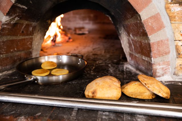 Close up detail of three calzone and a frying pan with buns on it lying in an oven made from bricks, with a fire in the background