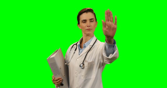 Female doctor holding medical folder while making a stop gesture with hand, set against a green screen background. Useful for themes around healthcare authority, medical advice, awareness campaigns, or healthcare presentations.