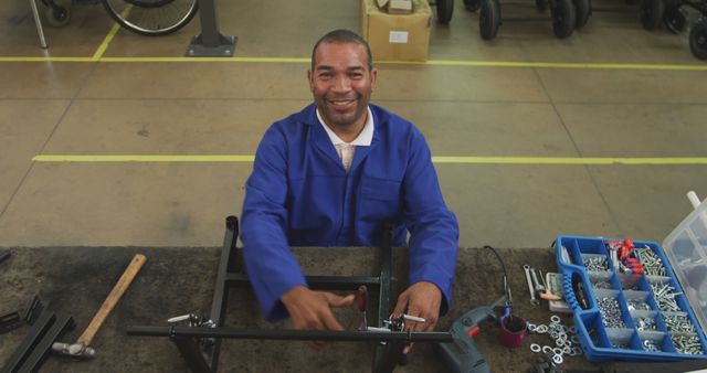 Afro-American technician working on mechanical assembly in factory environment, smiling at camera. Several tools and mechanical parts spread on workbench, showcasing engaged manual work and precision. Suitable for use in articles and advertisements related to engineering, manufacturing industry, industrial employment, machinery maintenance, and vocational training.