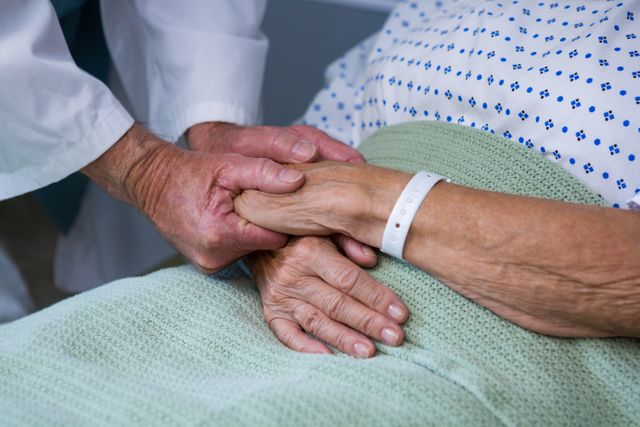 Doctor holding hand of senior patient lying in hospital bed, showing compassion and support. Useful for themes related to healthcare, elderly care, medical support, and patient-doctor relationships.