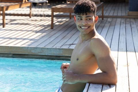 Smiling african american man relaxing on wooden pool deck in swim trunks under bright sun
