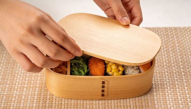 Hands lifting oval wooden bento box revealing rice corn seaweed salad and croquette lunch