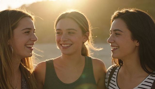 Three young women laughing on sunset beach, backlit golden glow, friendship moment