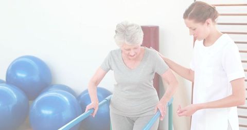 Senior woman walking between parallel bars while therapist assisting mobility