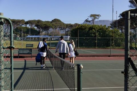Diverse tennis teammates walking across court carrying bags on sunny outdoor fenced court