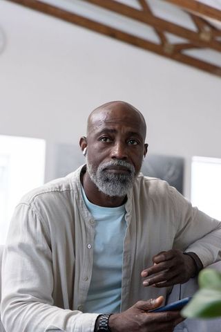 Thoughtful senior african man sitting and listening with earbud while holding smartphone
