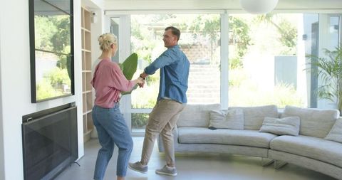 Sunlit couple dancing in modern living room near glass doors with curved sectional sofa