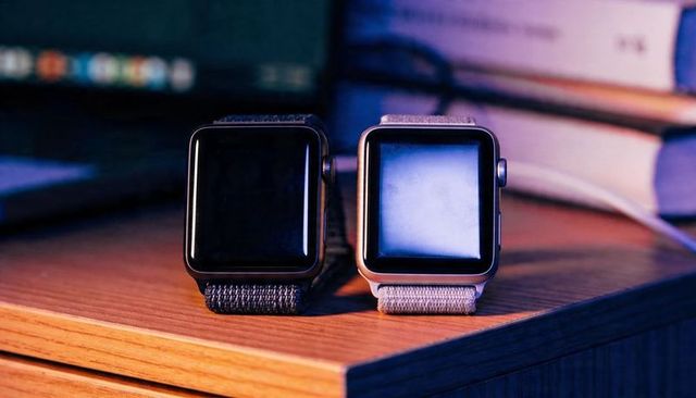 Pair of smartwatches with woven straps charging on wooden desk under ambient bokeh light