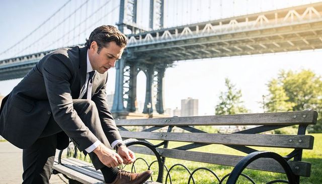 Businessman tying shoe on riverside bench beneath suspension bridge during morning commute