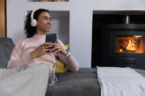 Nonbinary young adult relaxing on sofa listening with headphones by warm fireplace