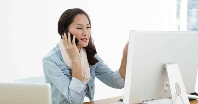 Young businesswoman wearing casual clothing talking on phone while using desktop computer in bright, modern office. Ideal for depicting professionalism, communication, technology use in business, multitasking ability. Suitable for business, corporate, technology, communication, and work-life balance themes.