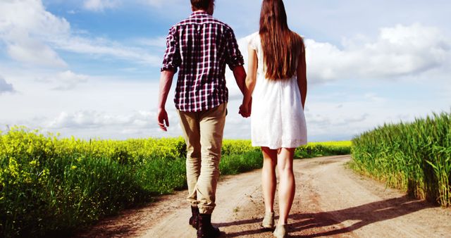 Young couple walking hand in hand along a dirt pathway between lush green and yellow fields under a partly cloudy blue sky. Useful for themes about romance, love, connection, outdoor activities, and serene landscapes. Can be used in blogs, articles, and advertisements related to relationships, travel, and lifestyle.