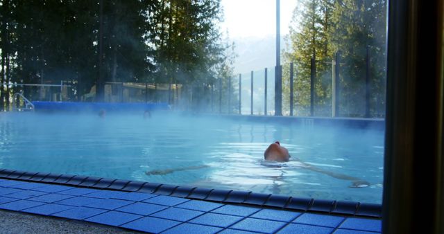 A person enjoys a swim in an outdoor pool with steam rising from the warm water on a chilly day, with copy space. The serene atmosphere suggests a tranquil retreat or a health and wellness setting.