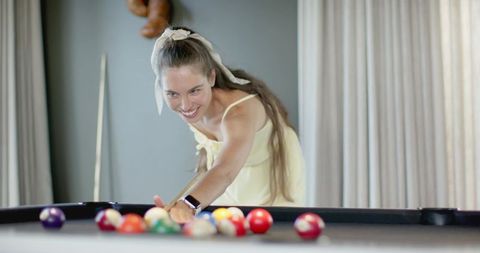 Woman leaning over pool table aiming cue while smiling wearing smartwatch and yellow dress