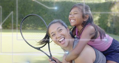 Mother daughter playing tennis piggyback, laughing while holding racket on sunlit court