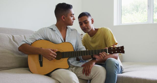 Smiling biracial gay male couple sitting on sofa and kissing while one plays guitar. staying at home in isolation during quarantine lockdown.
