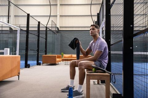 Male athlete resting at indoor padel court holding towel and racket, sports break