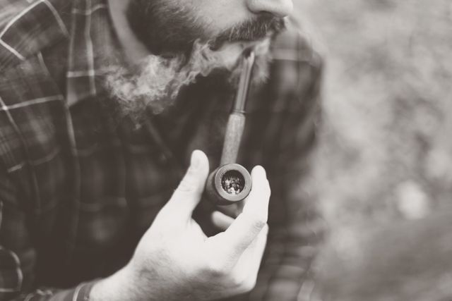 Man with beard smoking pipe outdoors in sepia tone. Perfect for use in articles on relaxation, vintage lifestyles, or contemplative moments. The close-up detail emphasizes the act of smoking, conveying a sense of leisure and nostalgia. Suitable for use in lifestyle magazines, blogs, or social media content related to traditional activities.